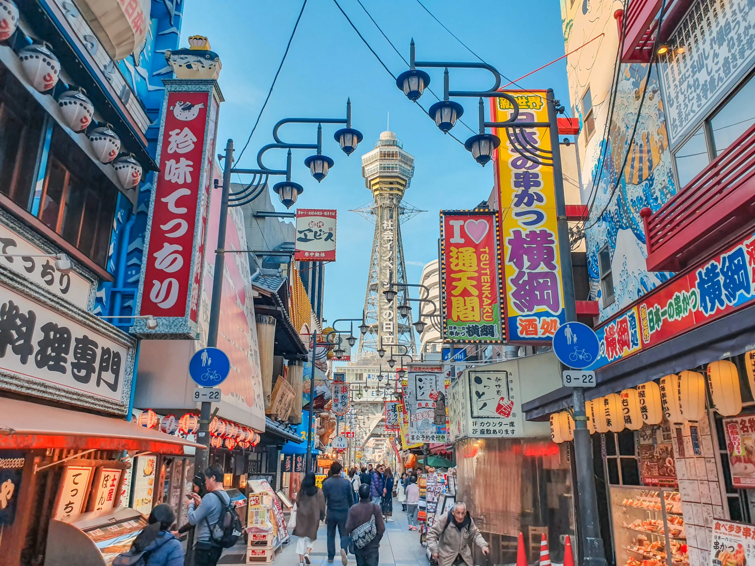 A bustling street in Osaka’s Shinsekai district, filled with colorful signs, shops, and Tsutenkaku Tower in the distance.