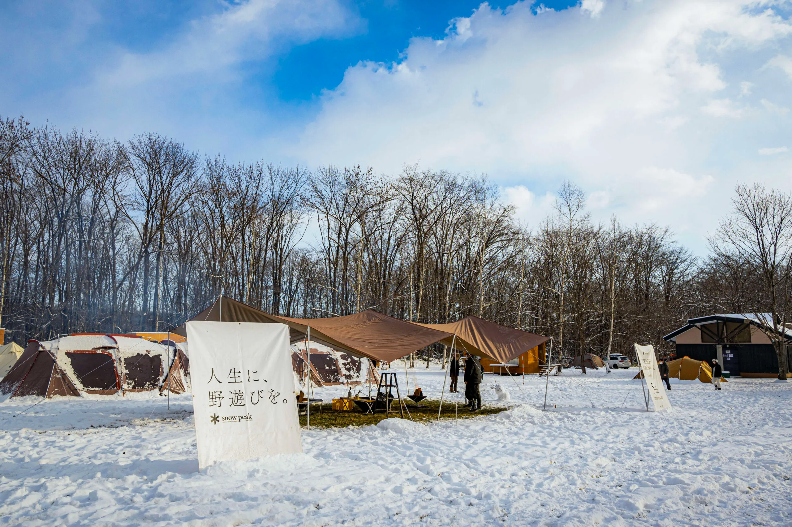 Snow-covered campground with tents and tarps set up under a clear winter sky, surrounded by bare trees.