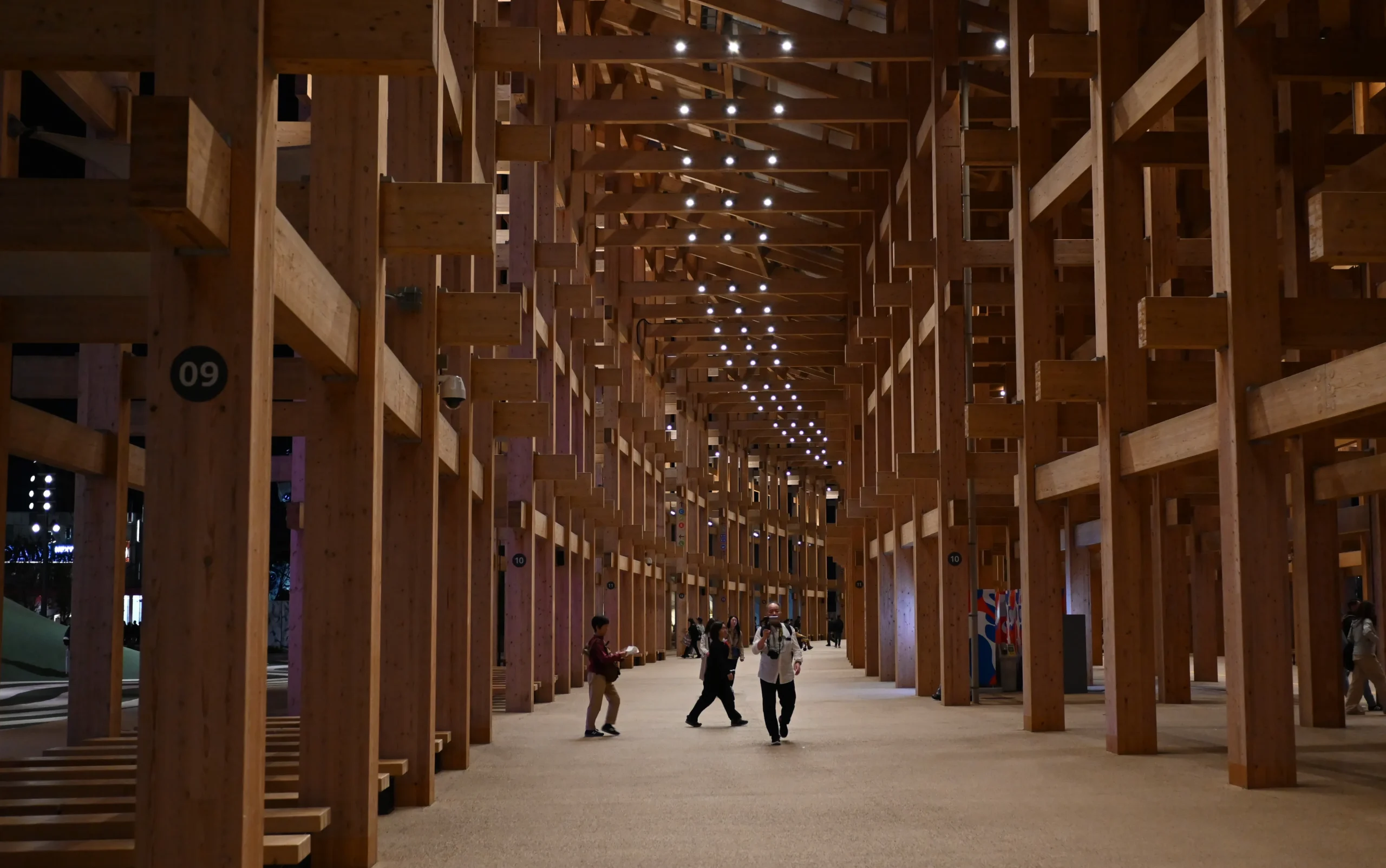 Visitors walk through a vast wooden structure with a grid of beams and ceiling lights at Expo 2025’s central ring.
