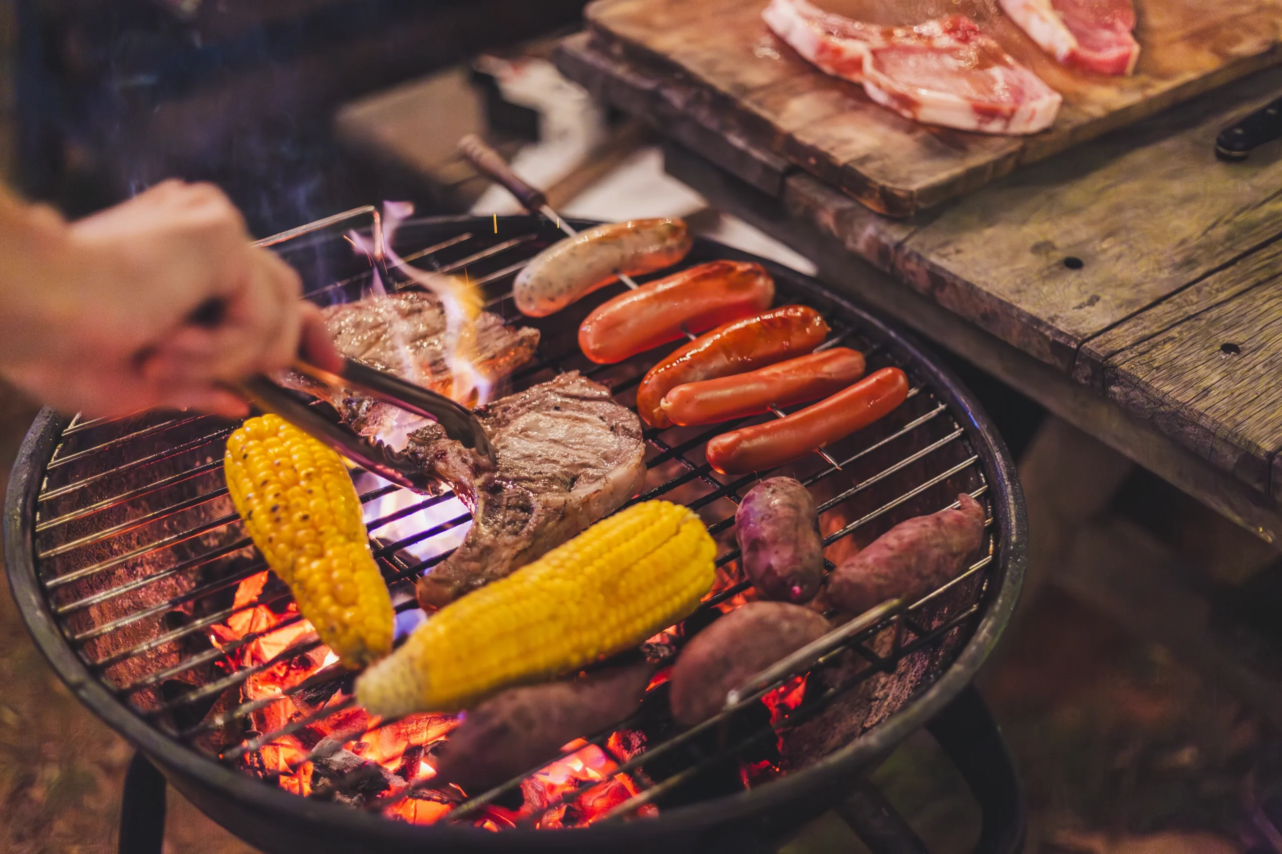 A close-up of a barbecue grill with sausages, corn, sweet potatoes, and steak cooking over open flames at a campsite.