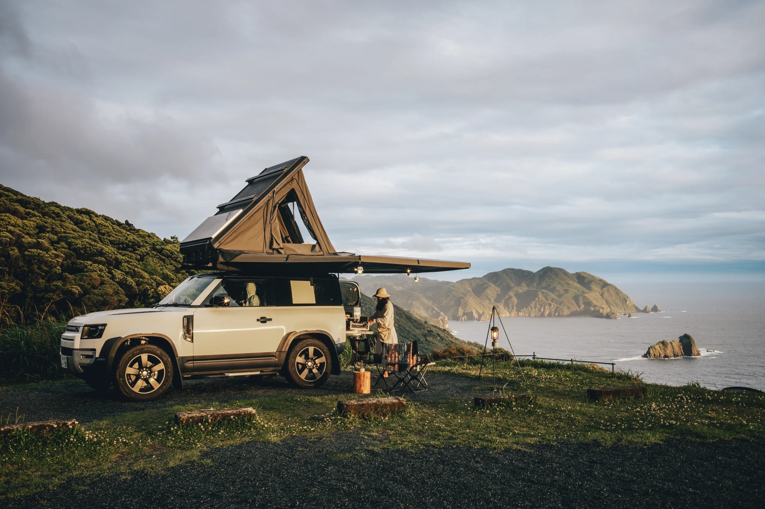 A rooftop tent setup on a Land Rover parked at a scenic coastal viewpoint in Japan, with a camper preparing a meal.