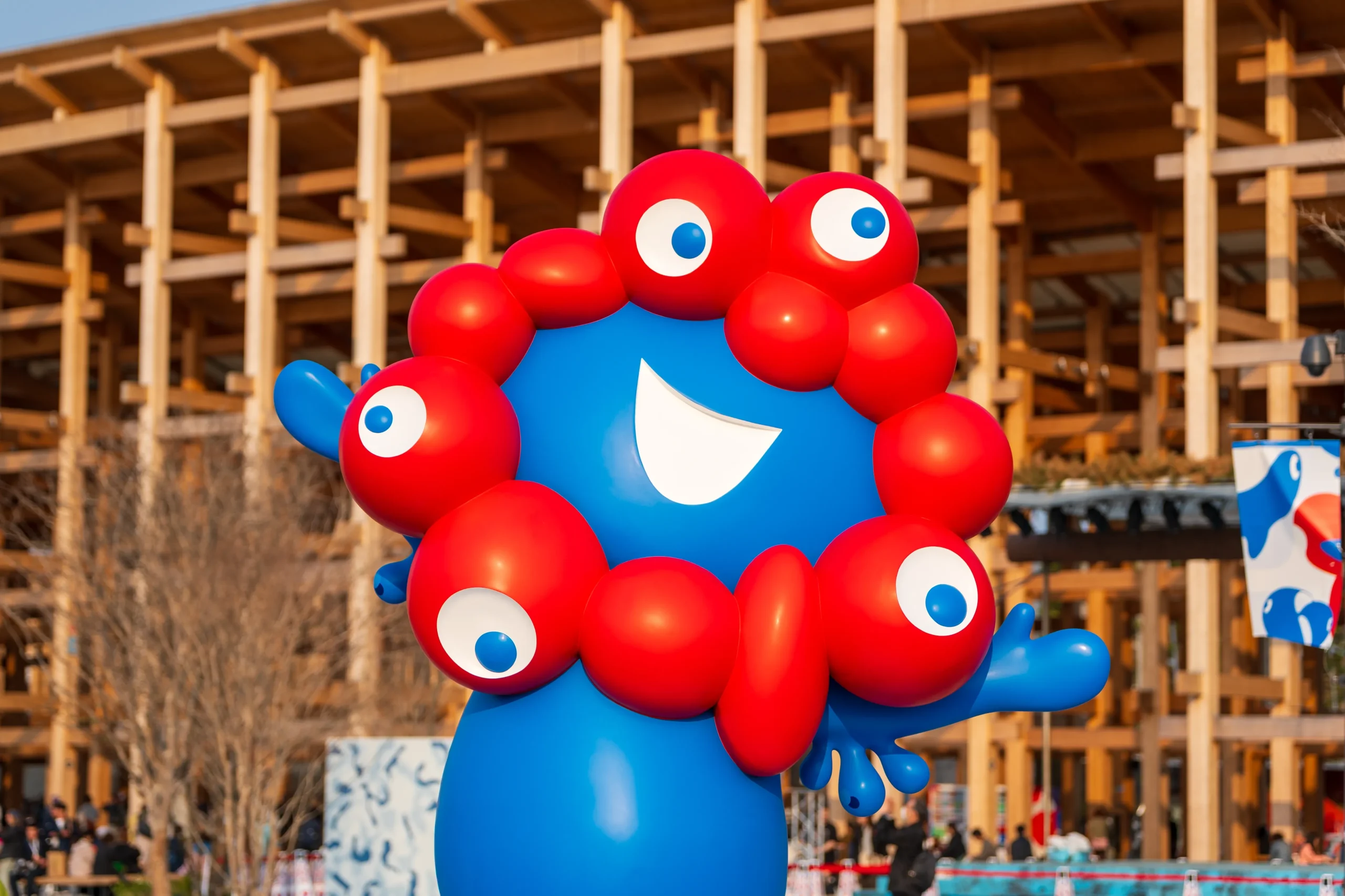 Large, colorful statue of Myaku-Myaku, the Expo 2025 mascot, smiling under a blue sky with a crowd in the background.