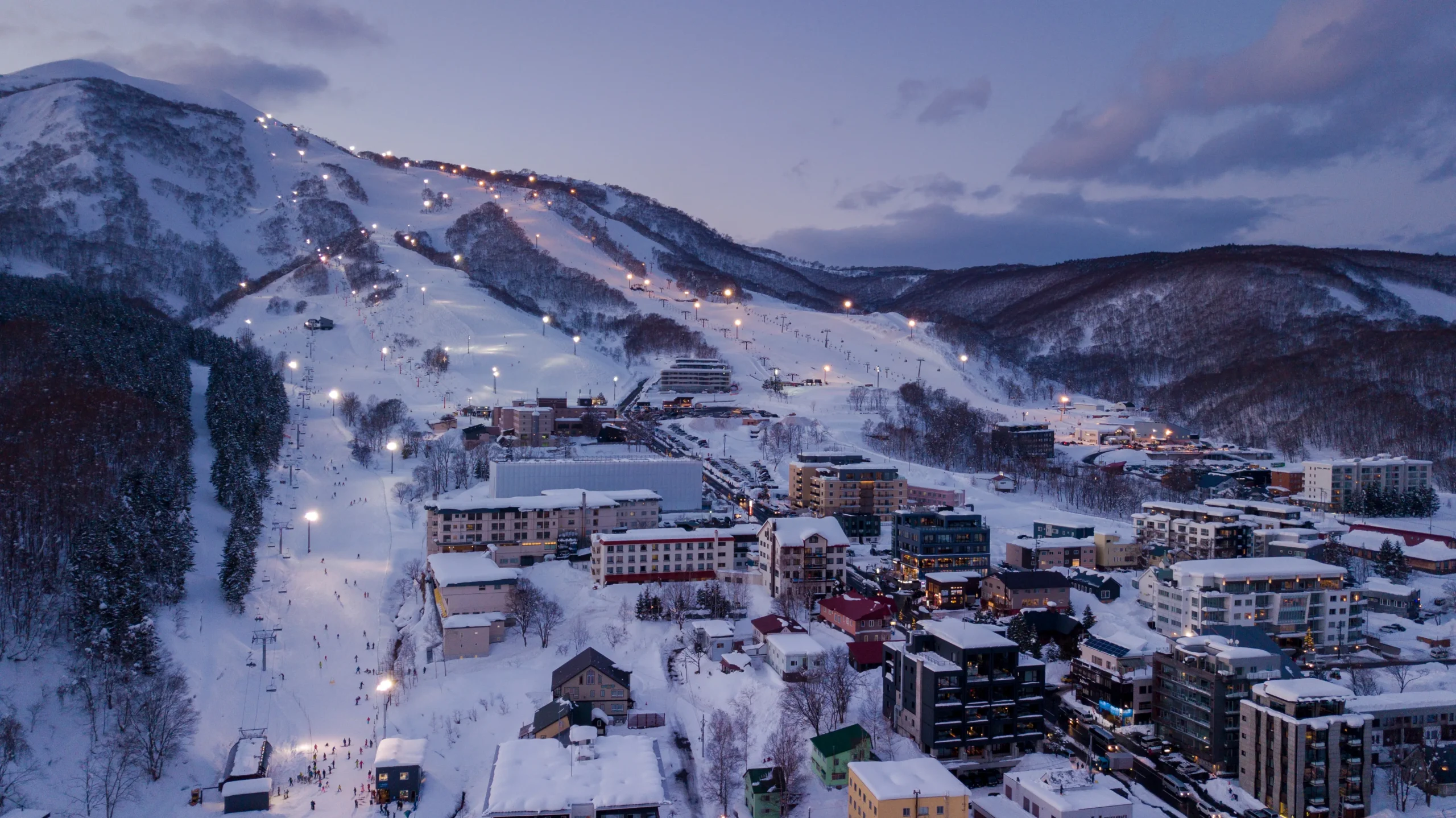 Aerial view of Niseko’s illuminated ski slopes and alpine village during twilight, surrounded by snow-covered mountains.