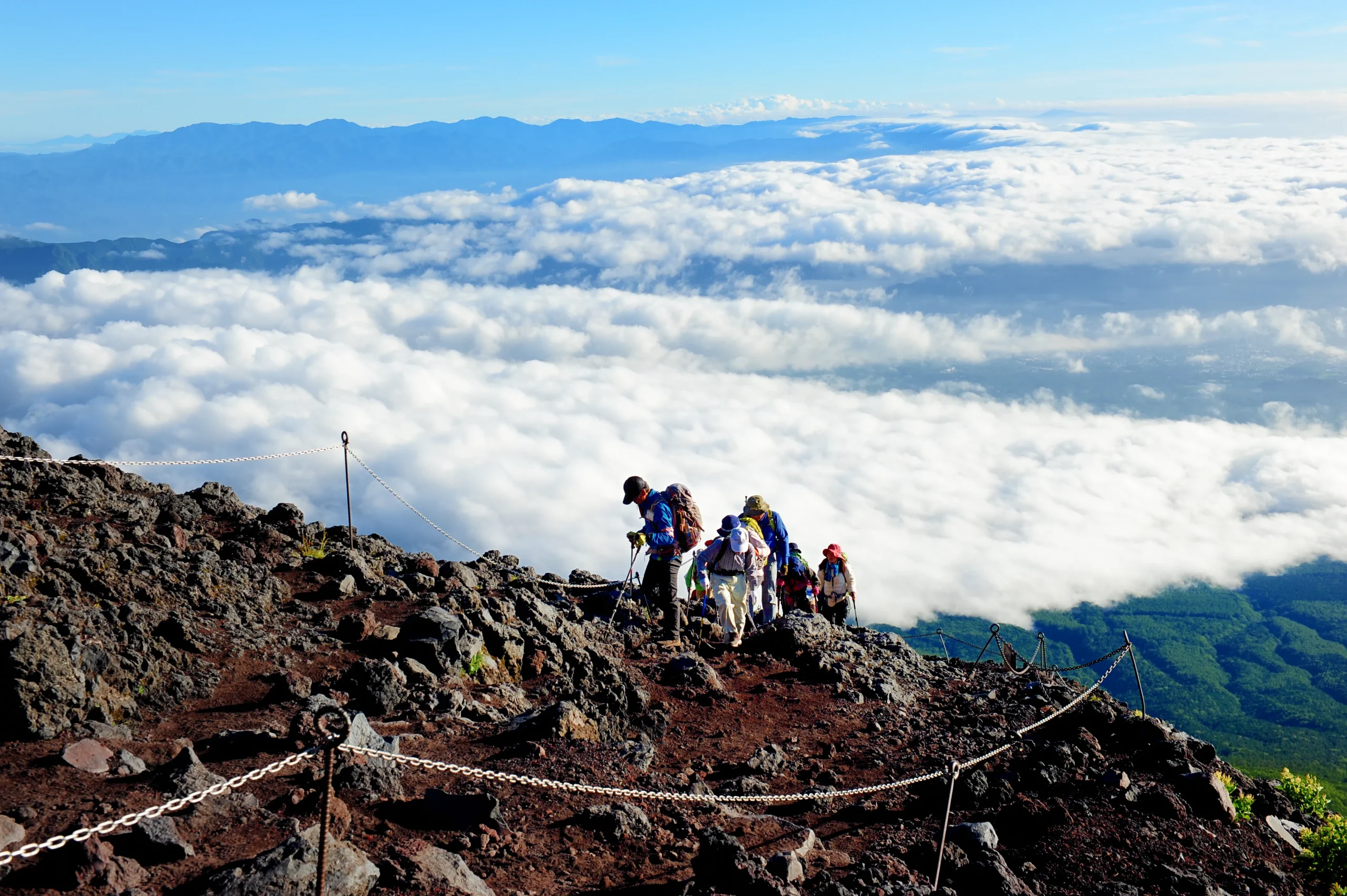 Hikers climbing a rocky trail on Mt. Fuji above a sea of clouds in Japan.