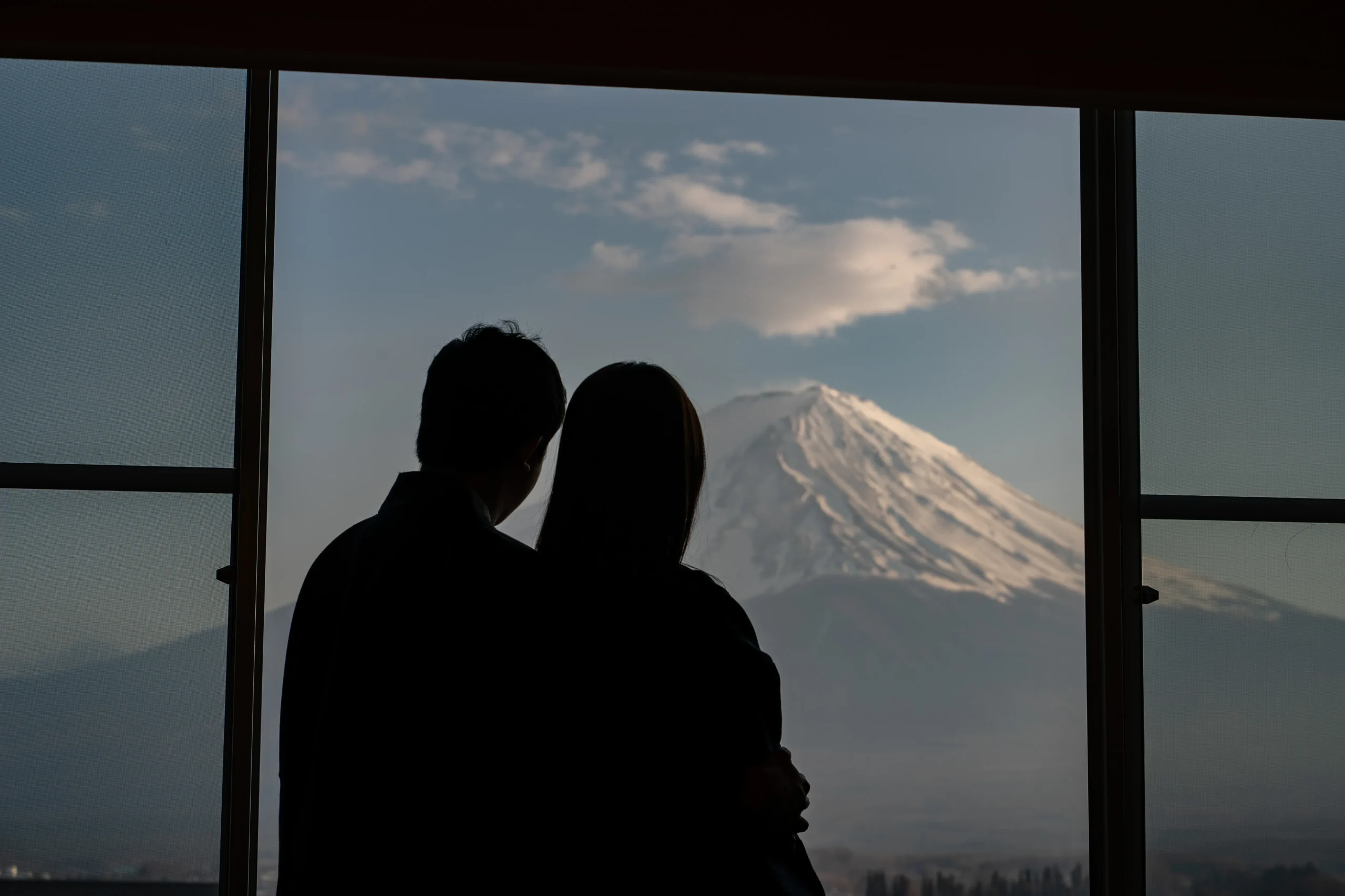 Silhouette of a couple at a ryokan looking at a snow-capped Mt. Fuji through a window.