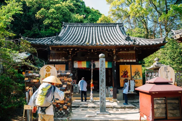 A historic Buddhist temple along the Shikoku 88 Temple Pilgrimage route, with an intricately carved wooden roof, colorful ema plaques, and visitors in pilgrimage attire.