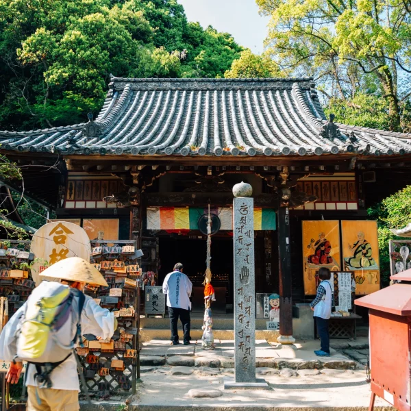 A historic Buddhist temple along the Shikoku 88 Temple Pilgrimage route, with an intricately carved wooden roof, colorful ema plaques, and visitors in pilgrimage attire.