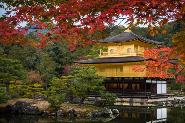 The Golden Pavilion in Kyoto, reflecting in a calm pond and framed by brilliant red autumn leaves and manicured trees.