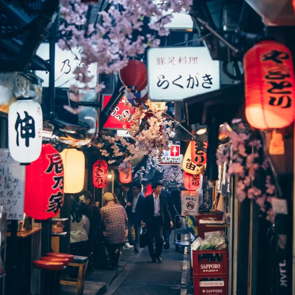 A narrow alley in Omoide Yokocho, Tokyo, lined with izakayas illuminated by red and white paper lanterns, with cherry blossoms adding to the nostalgic atmosphere.