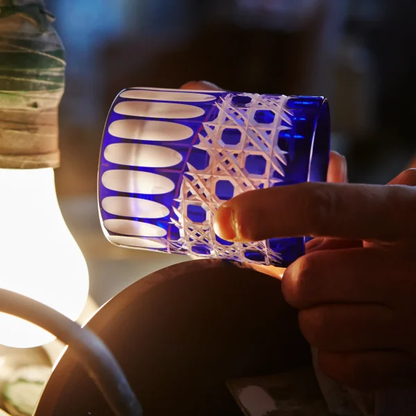 A craftsman carefully refining the edges of an Edo Kiriko glass, holding it against a rotating grinding wheel under the glow of a work lamp.