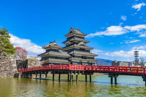 Historic Matsumoto Castle in Nagano reflected across the moat, with its iconic black keep and bright red bridge under a clear blue sky