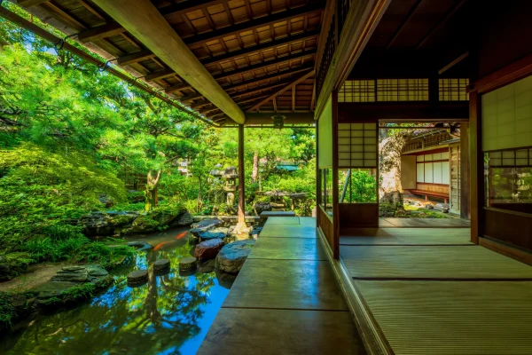 A traditional Japanese garden seen from the wooden veranda of a historical residence, overlooking a koi pond, stepping stones, and lush greenery.