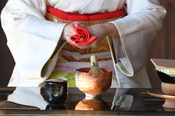 A woman dressed in a white kimono with a red obi performing a Japanese tea ceremony, delicately holding a folded red cloth, with a tea whisk and ceramic bowl on the table.