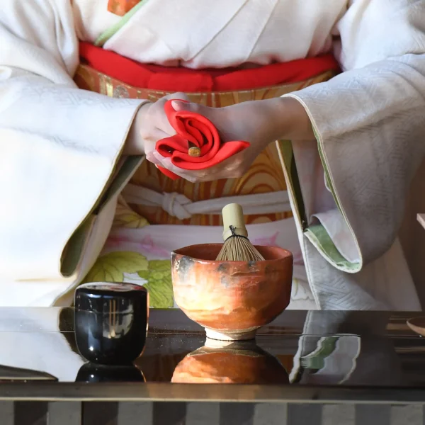 A woman dressed in a white kimono with a red obi performing a Japanese tea ceremony, delicately holding a folded red cloth, with a tea whisk and ceramic bowl on the table.